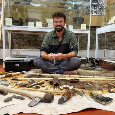 A man sits on the floor with a range of replica prehistoric tools laid out on animal skins in front of him. In the background are cabinets of some of thelater Prehistoric artefacts at the Museum of Islay Life