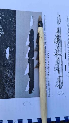 A replica mesolithic arrow is laid next to an illustration of an archaeological find from southern Sweden. The arrow has a flint microlith tip & four triangular microlith barbs set in a groove along the shaft.